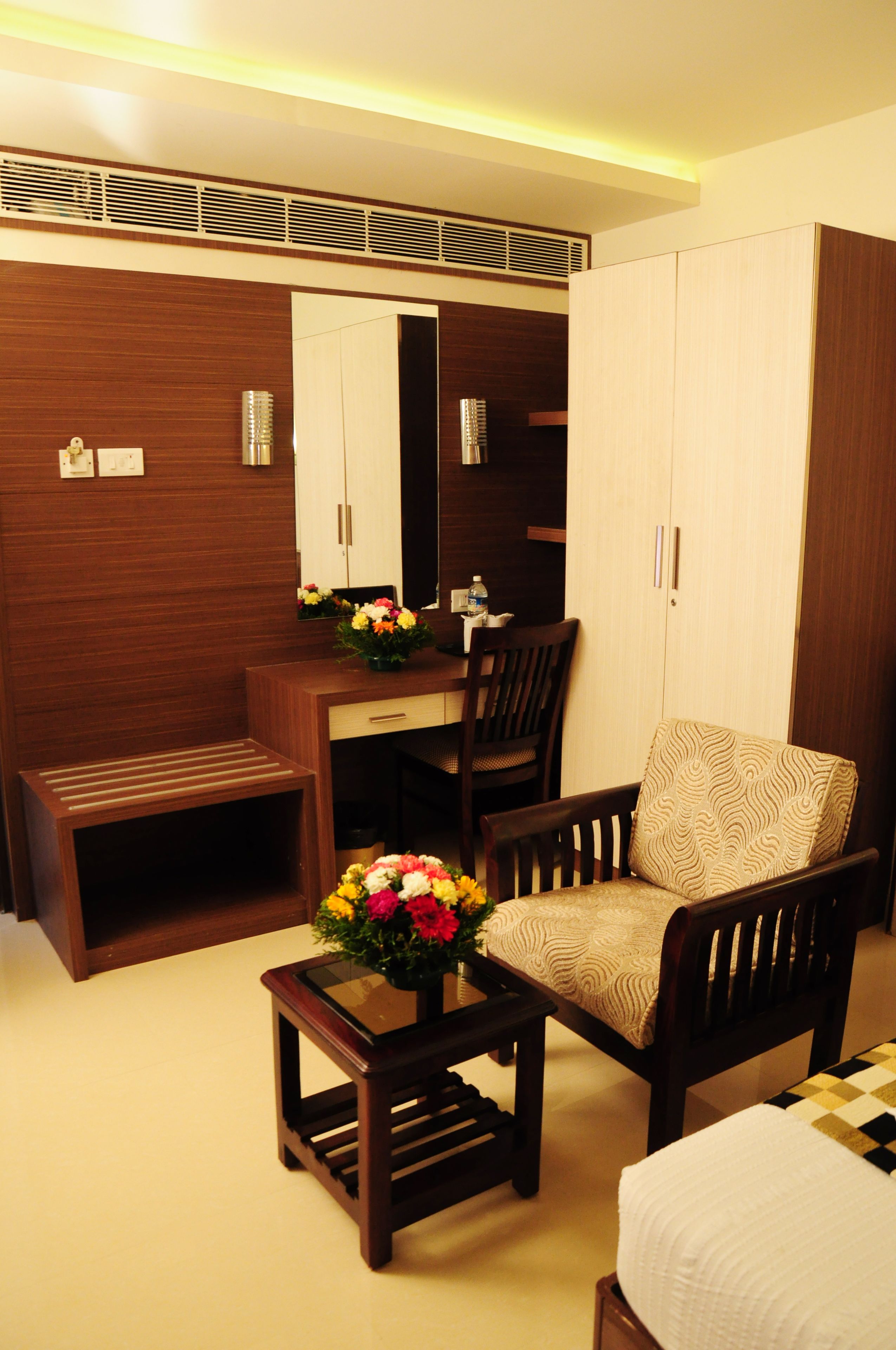 A hotel room with a wooden desk, a chair, a small table with a vase of flowers, a wardrobe, and a bed.
