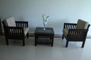 Two dark wooden chairs with patterned cushions, a small square table with a vase of white flowers, and a plain wall in the background.