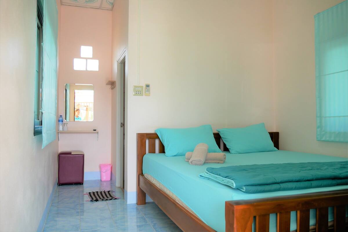 A bedroom with a bed, a suitcase, a pink trash bin, and a window with natural light.