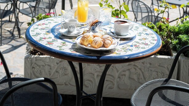 A round table with a floral tablecloth, a plate of pastries, a cup, a glass of orange juice, and a small potted plant. - Villa Amore (Ravello)
