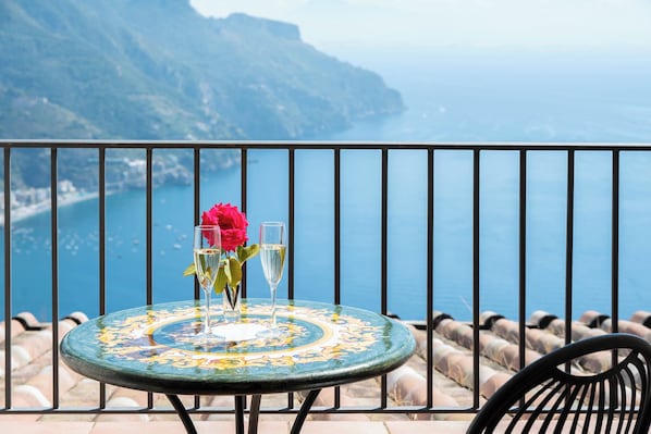 A round table with a floral pattern, a glass of champagne, and a red rose on a balcony overlooking a coastal view.