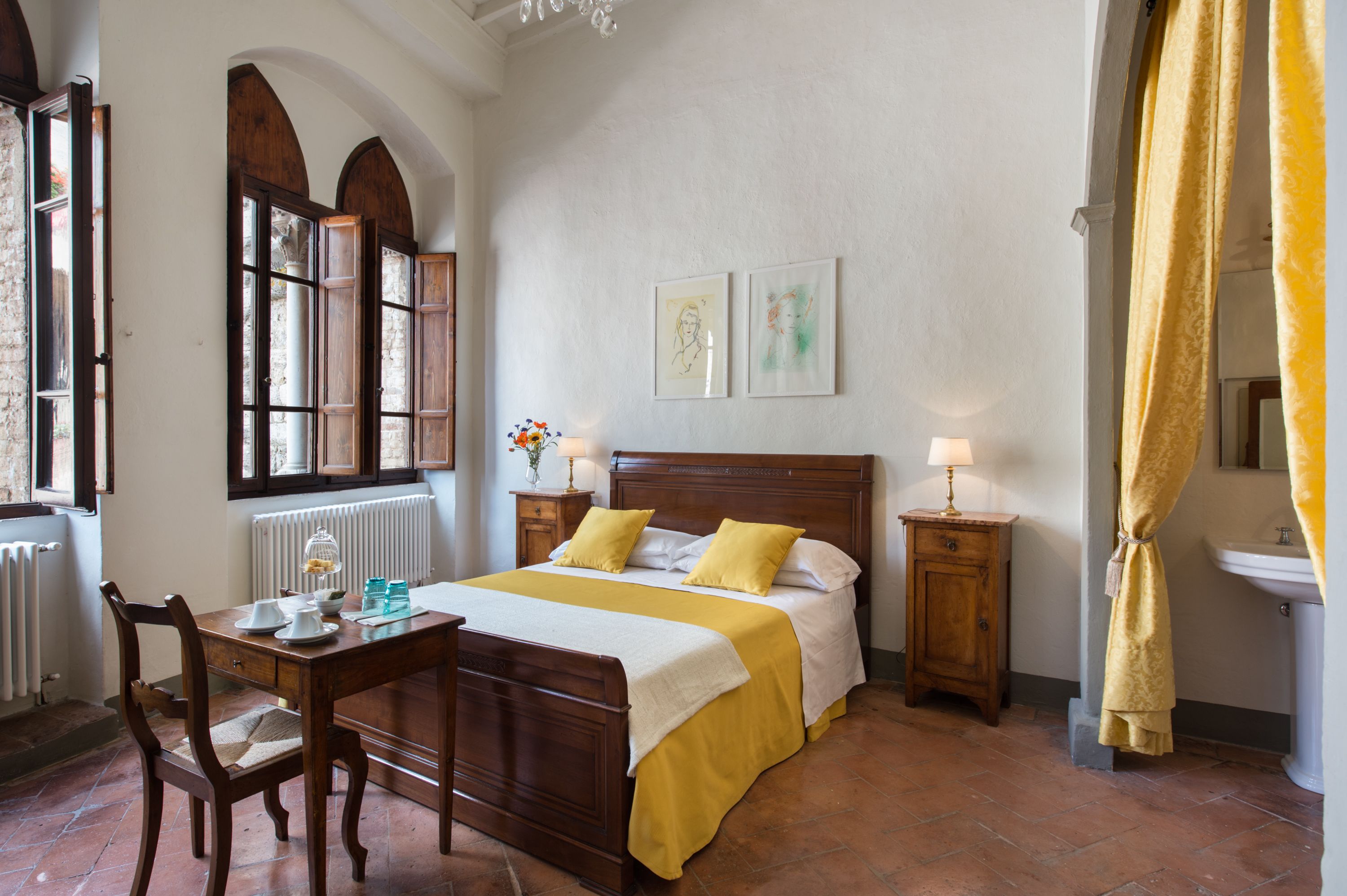 A bedroom with a wooden bed, a bedside table with a lamp, a chair, a window with wooden shutters, and a radiator.