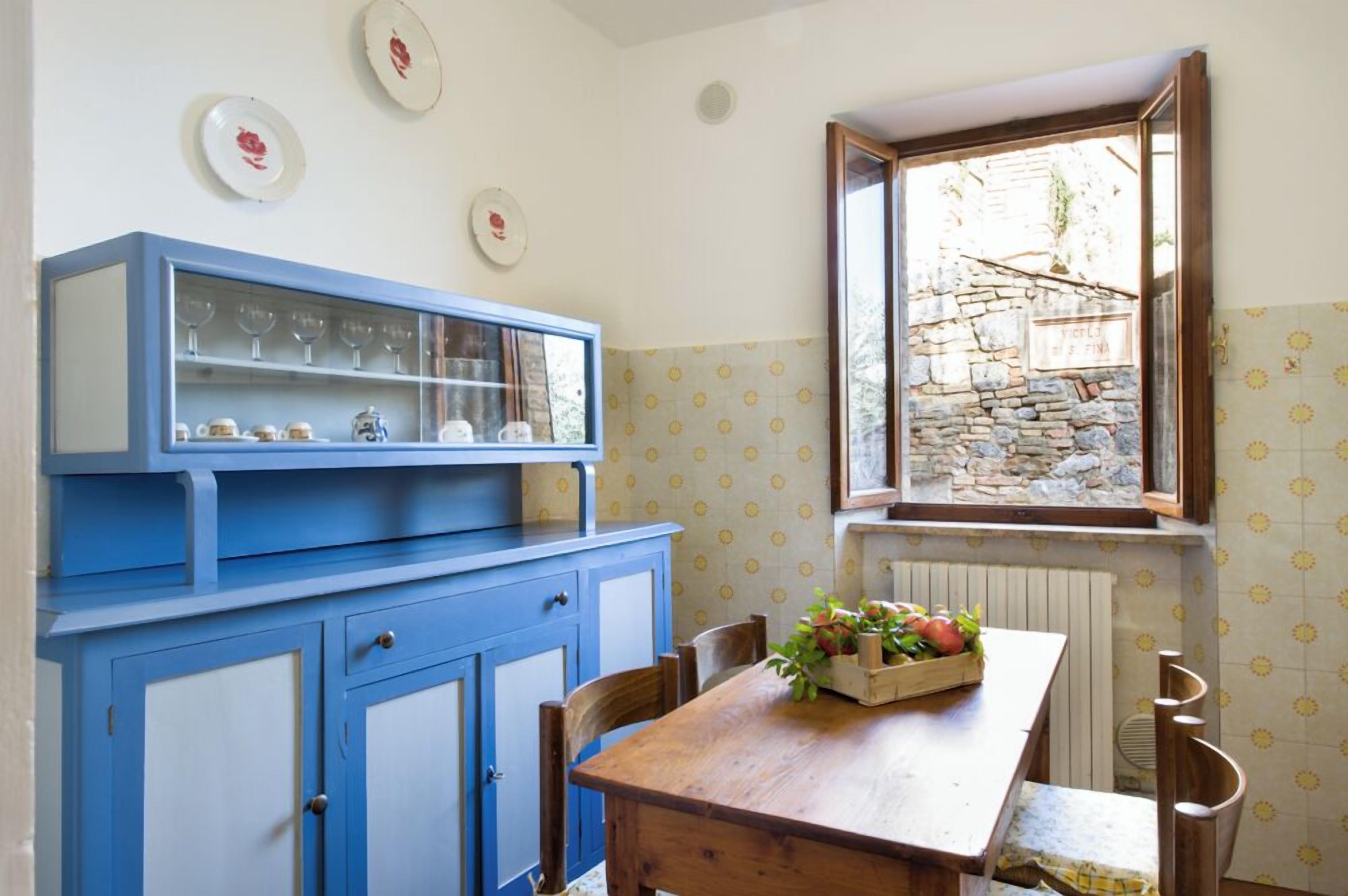 A kitchen with blue cabinets, a wooden table, and a window with a stone wall view.