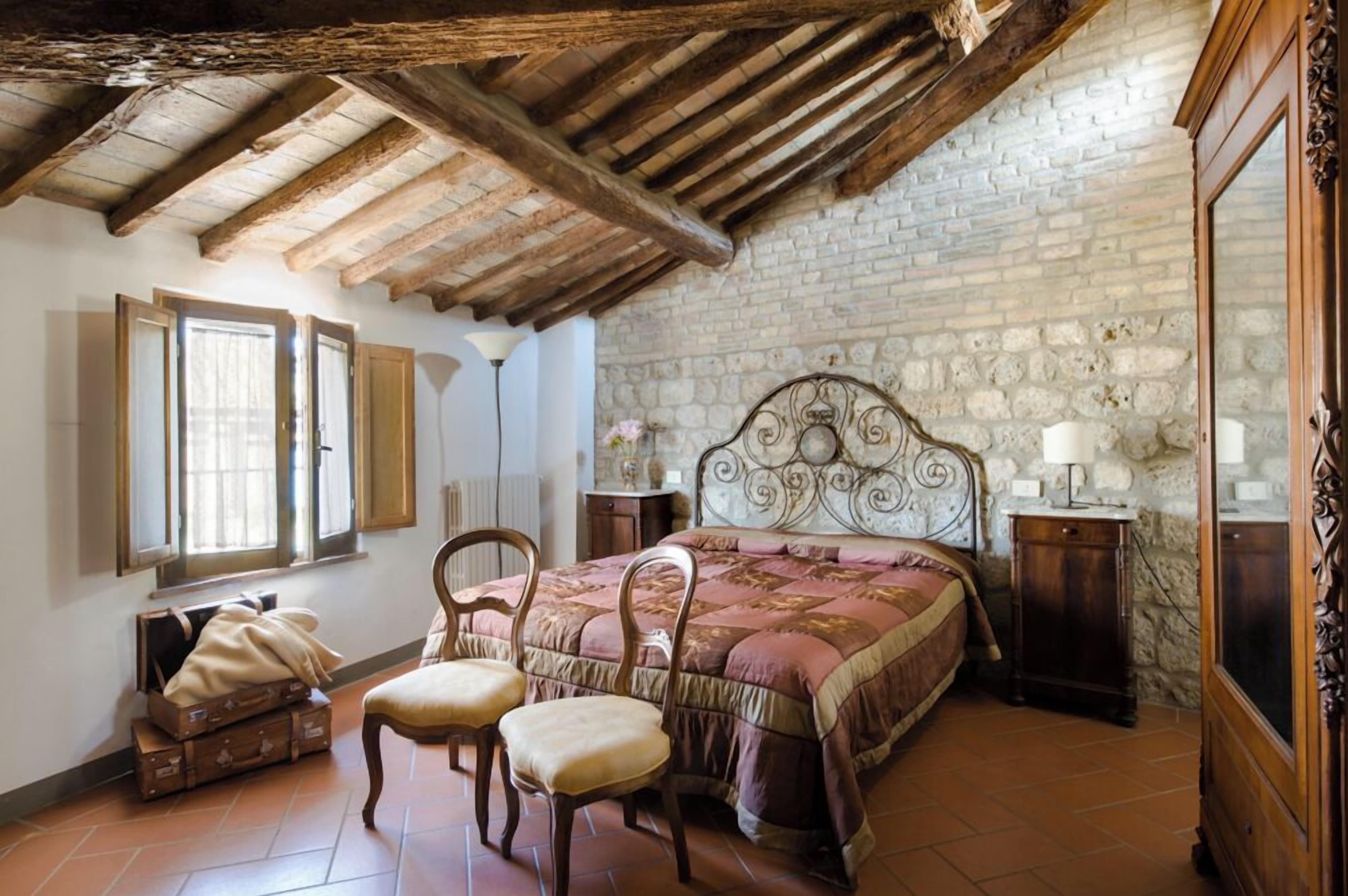 Un dormitorio con pared de piedra rústica, vigas de madera en el techo, una cama con cabecera decorativa, dos sillas, una mesita de noche con lámpara y una ventana con contraventanas.