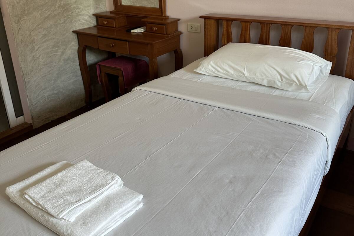 Ein ordentlich bezogenes Bett mit weißen Laken und einem gefalteten Handtuch, ein Nachttisch aus Holz mit einem Spiegel und ein Holz-Kopfstück.