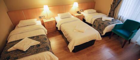 A hotel room with three single beds, each with a bedside table and lamp, a green chair, and a wooden floor.