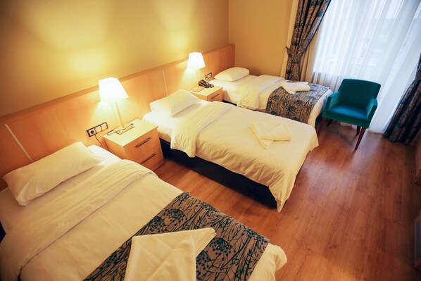 A hotel room with three beds, a desk with a lamp, a chair, and a window with curtains.
