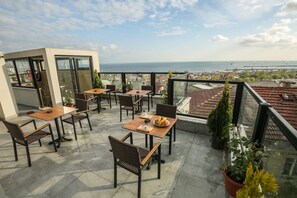 A terrace with tables and chairs, potted plants, and a view of the sea and cityscape.