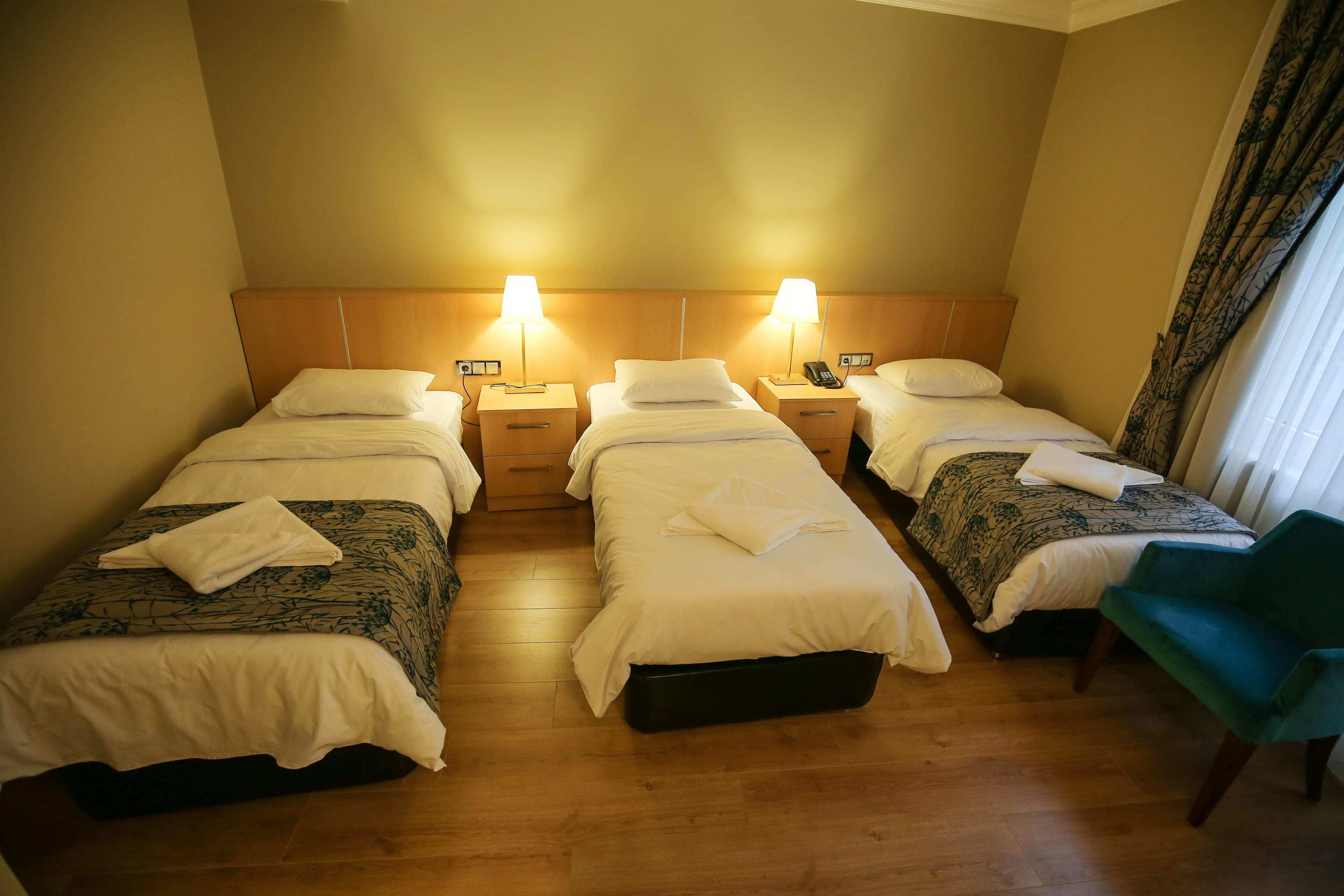 A hotel room with three single beds, each with a bedside table and lamp, and a green chair.
