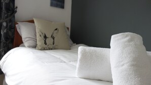 Un lit bien fait, avec du linge de lit blanc et une serviette blanche, un oreiller orné d’un motif noir et un mur sombre en arrière-plan.