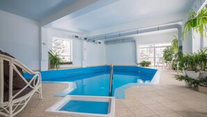 Una gran piscina interior con agua cristalina azul, rodeada de plantas en macetas y una silla de mimbre.