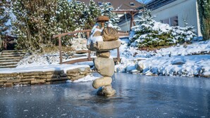 Un equilibrio de piedras sobre una superficie helada, con una casa y árboles cubiertos de nieve al fondo.