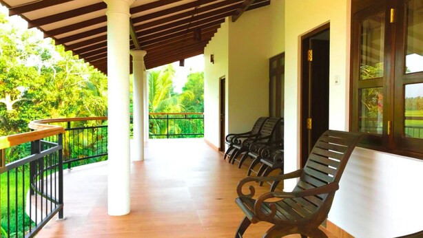 A covered outdoor area with wooden flooring, white pillars, and black metal railings. There are several black chairs arranged along the wall. - Lavender Home Rest (Thissamaharama)