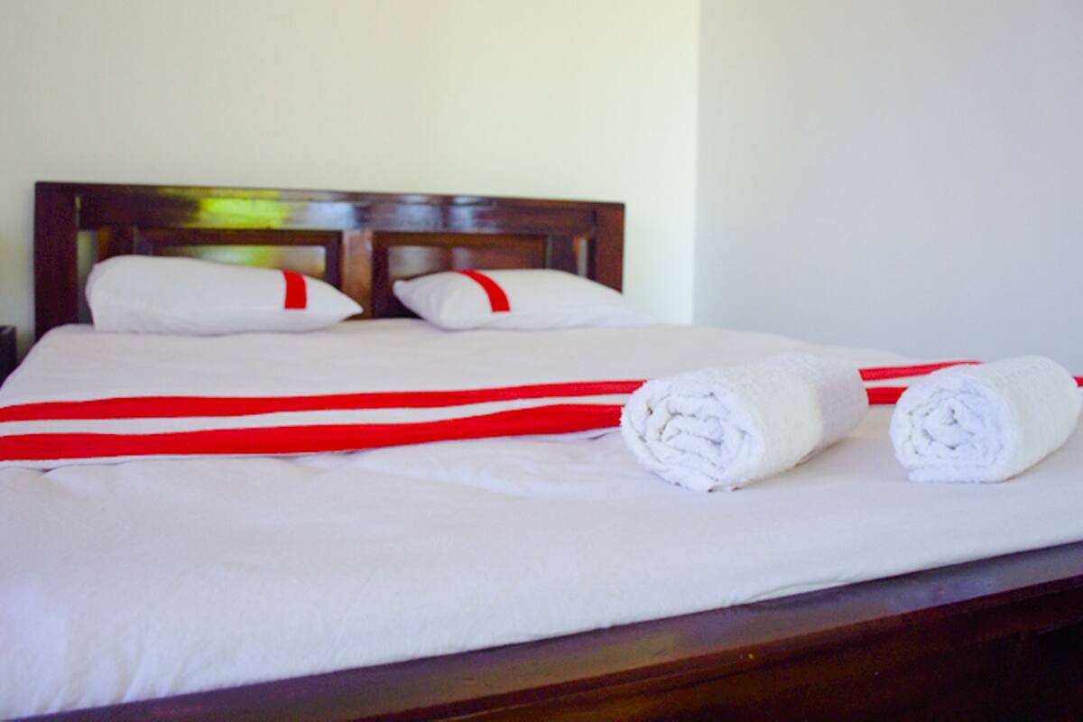 A neatly made bed with white linens and red accents, a wooden headboard, and two rolled towels on the bed.