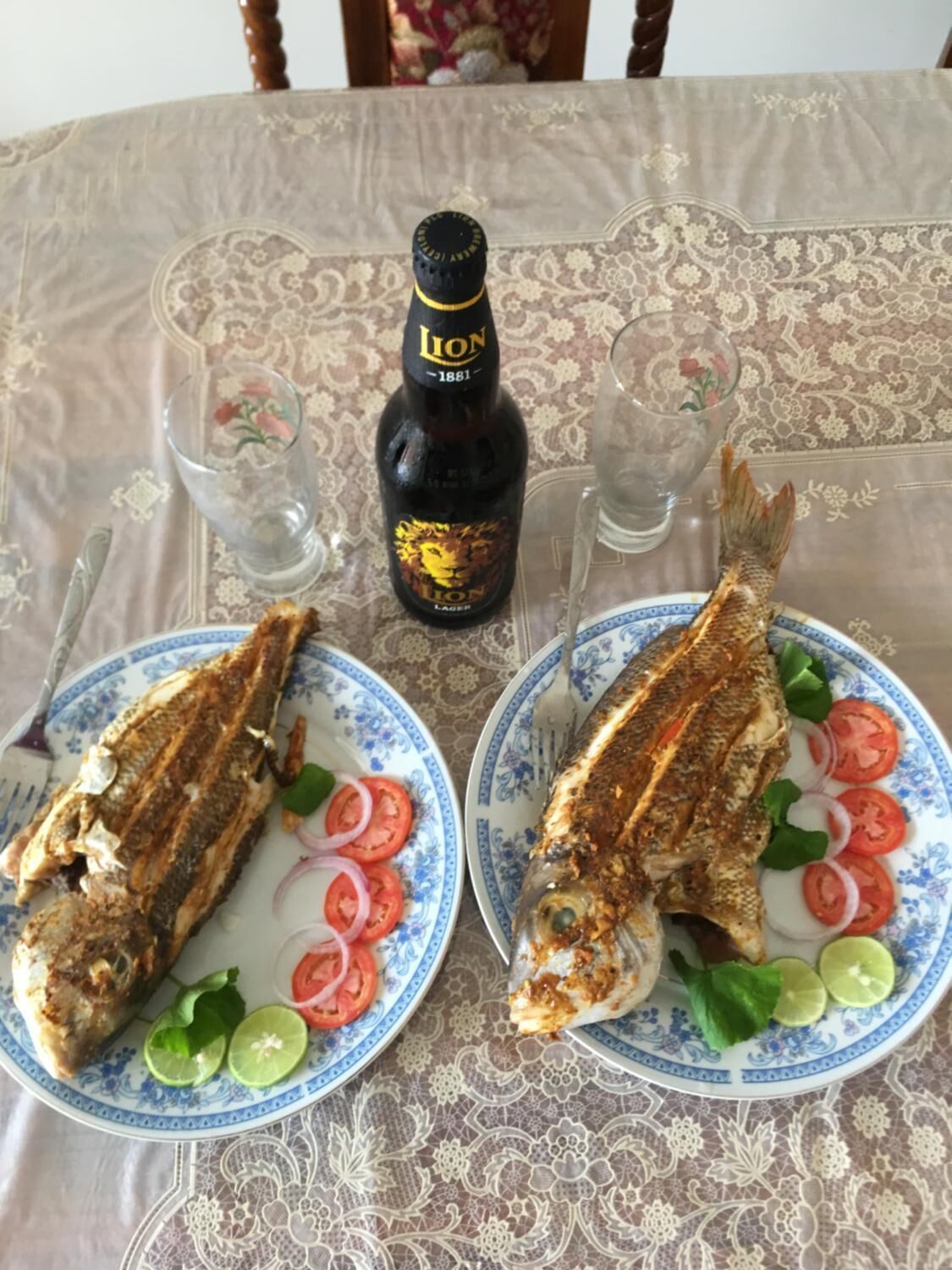Two plates of grilled fish with lemon slices, tomatoes, and onions, a bottle of Lion beer, and two glasses on a table with a floral tablecloth.