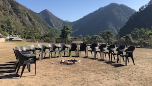 A seating area with plastic chairs arranged in a semi-circle around a fire pit, set against a backdrop of mountains and a clear sky. - Ecoterra Solace Resort (Narendranagar)