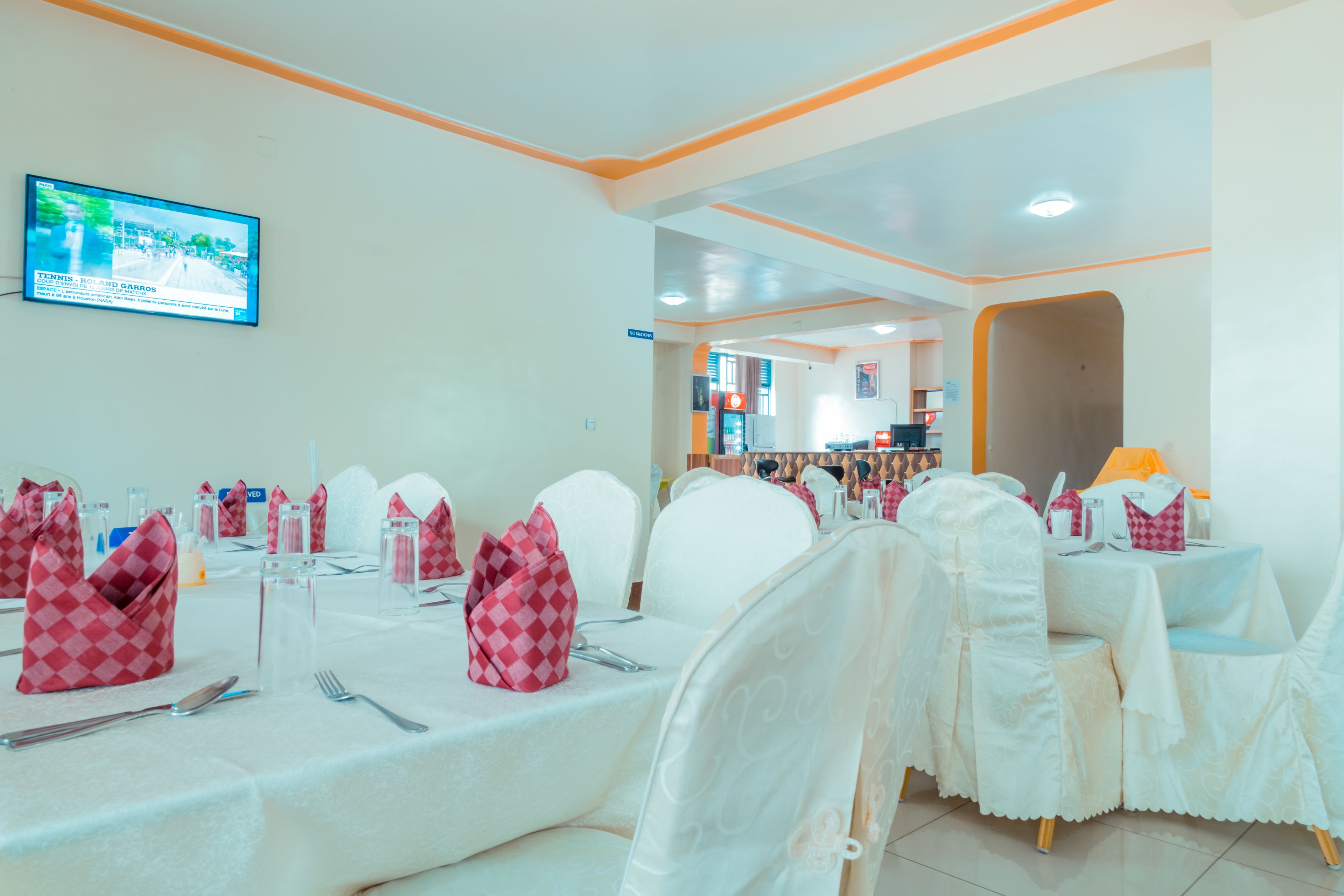 A dining area with tables set for a meal, chairs with white covers, and a television mounted on the wall displaying a tennis match.
