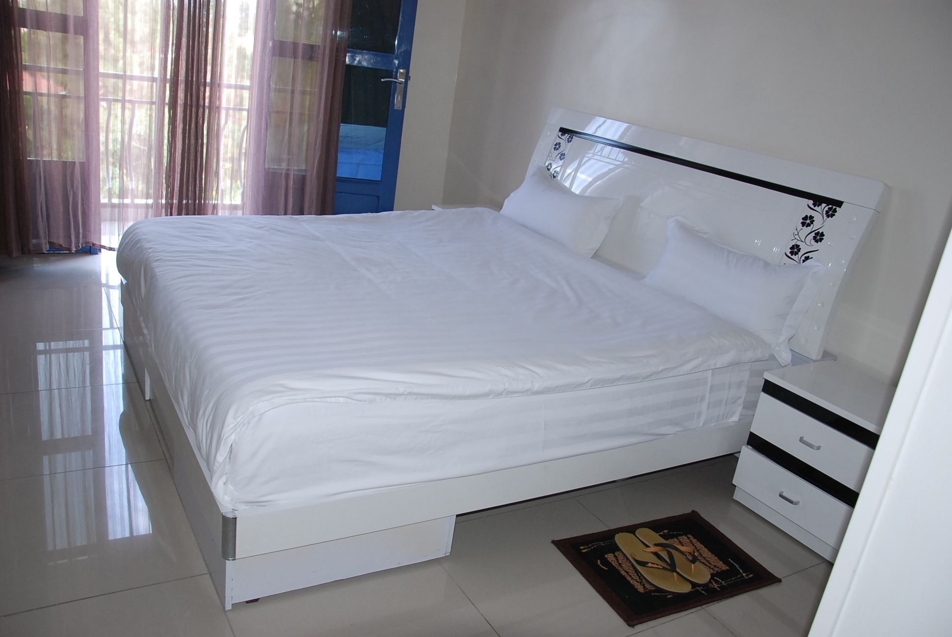 A neatly made bed with white linens in a room with a blue door and a white nightstand with drawers.