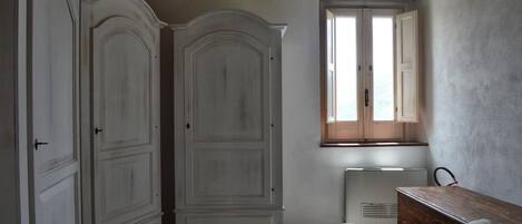 A room with a large white wardrobe, a small table, and a window with wooden shutters.