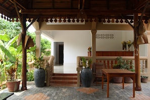 A covered outdoor area with a wooden table and chairs, potted plants, and a view of a building in the background.