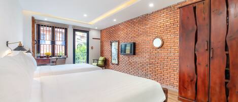A bedroom with a large bed, a wooden wardrobe, a TV mounted on the brick wall, and a desk with a chair.