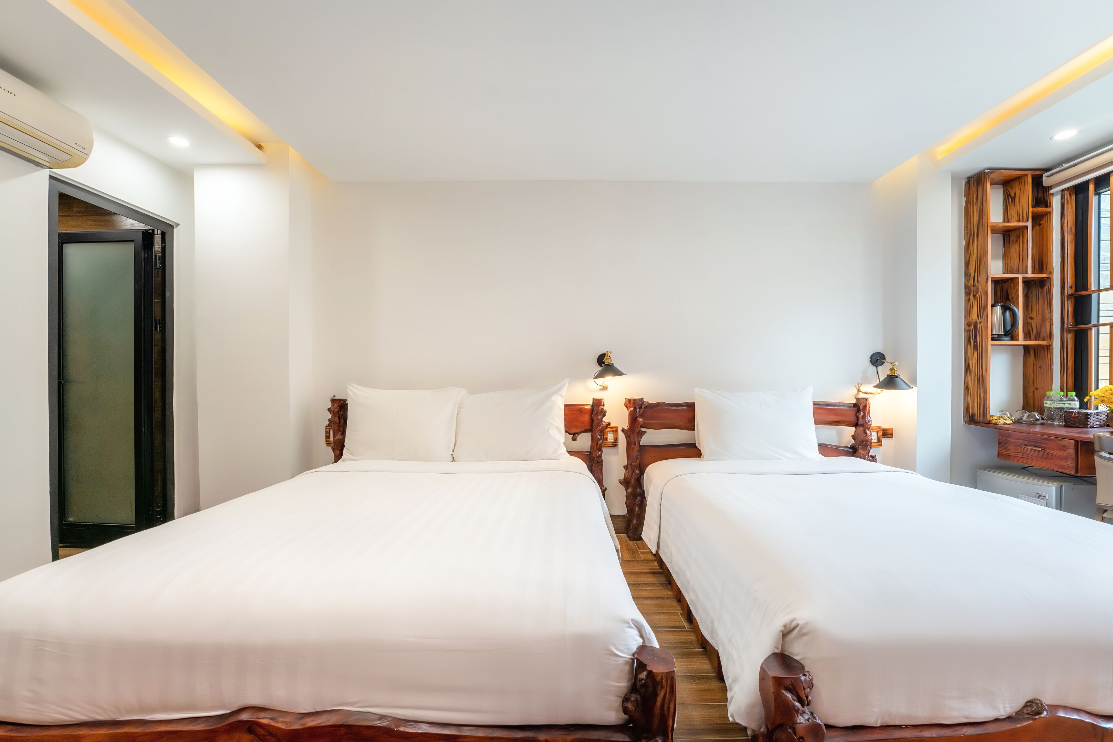 Une chambre d’hôtel avec deux lits simples, des têtes de lit en bois, un petit bureau avec une lampe, un miroir et une étagère en bois ornée d’objets décoratifs.