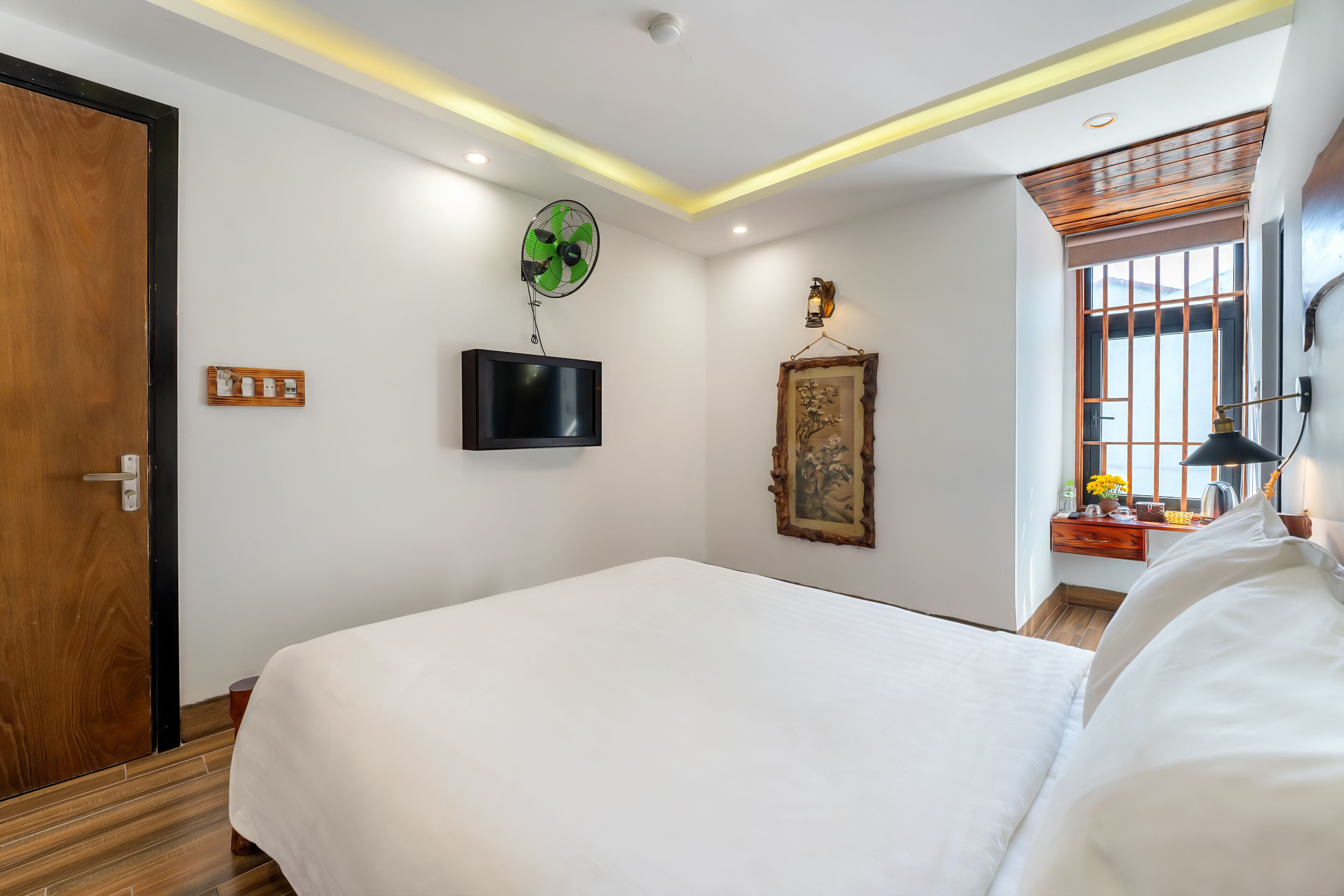 Habitación de hotel con una cama grande, un televisor montado en la pared, un ventilador de techo, un cuadro enmarcado y una ventana con barrotes.