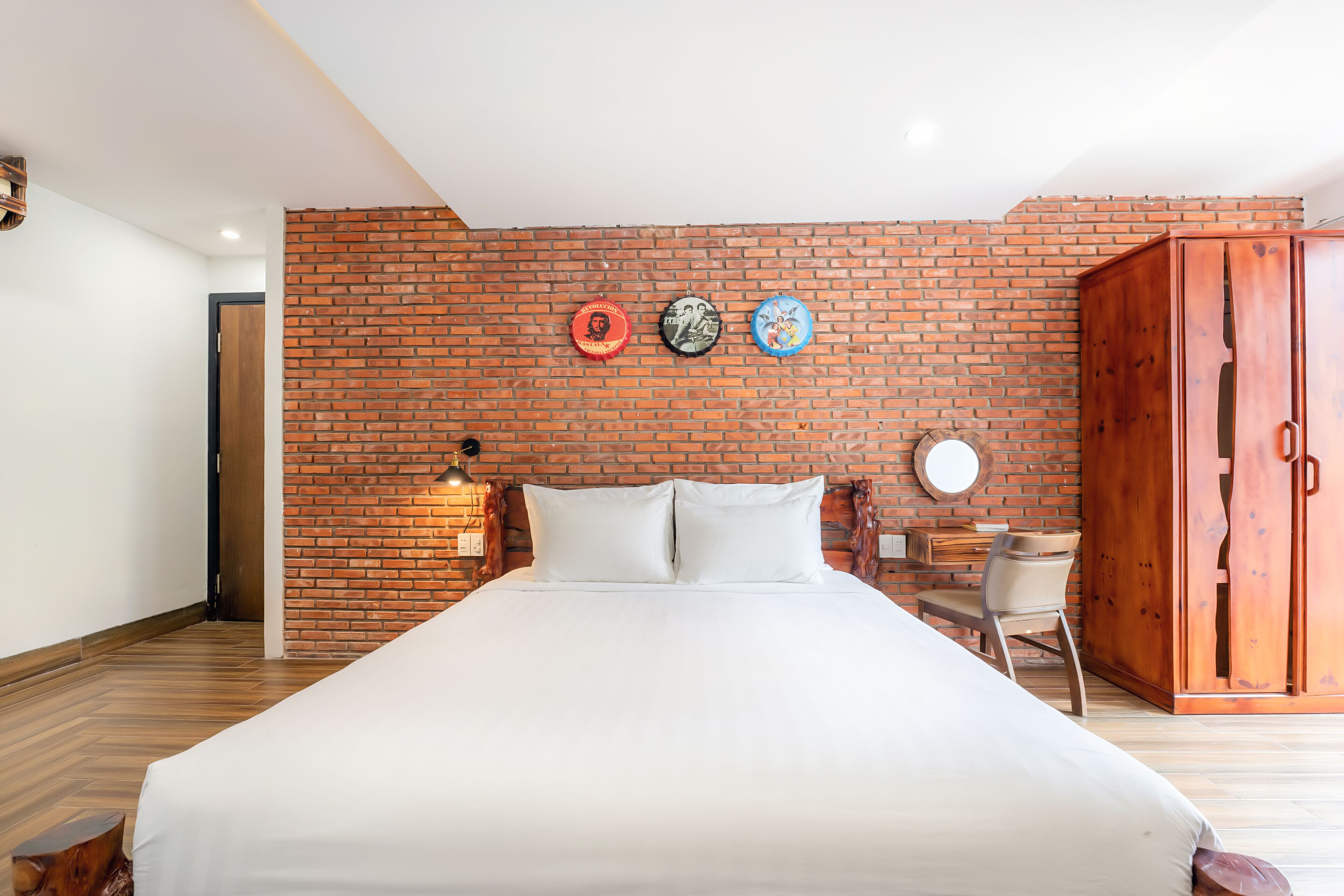 A bedroom with a brick wall, a bed with white bedding, a wooden wardrobe, and a small desk with a chair.
