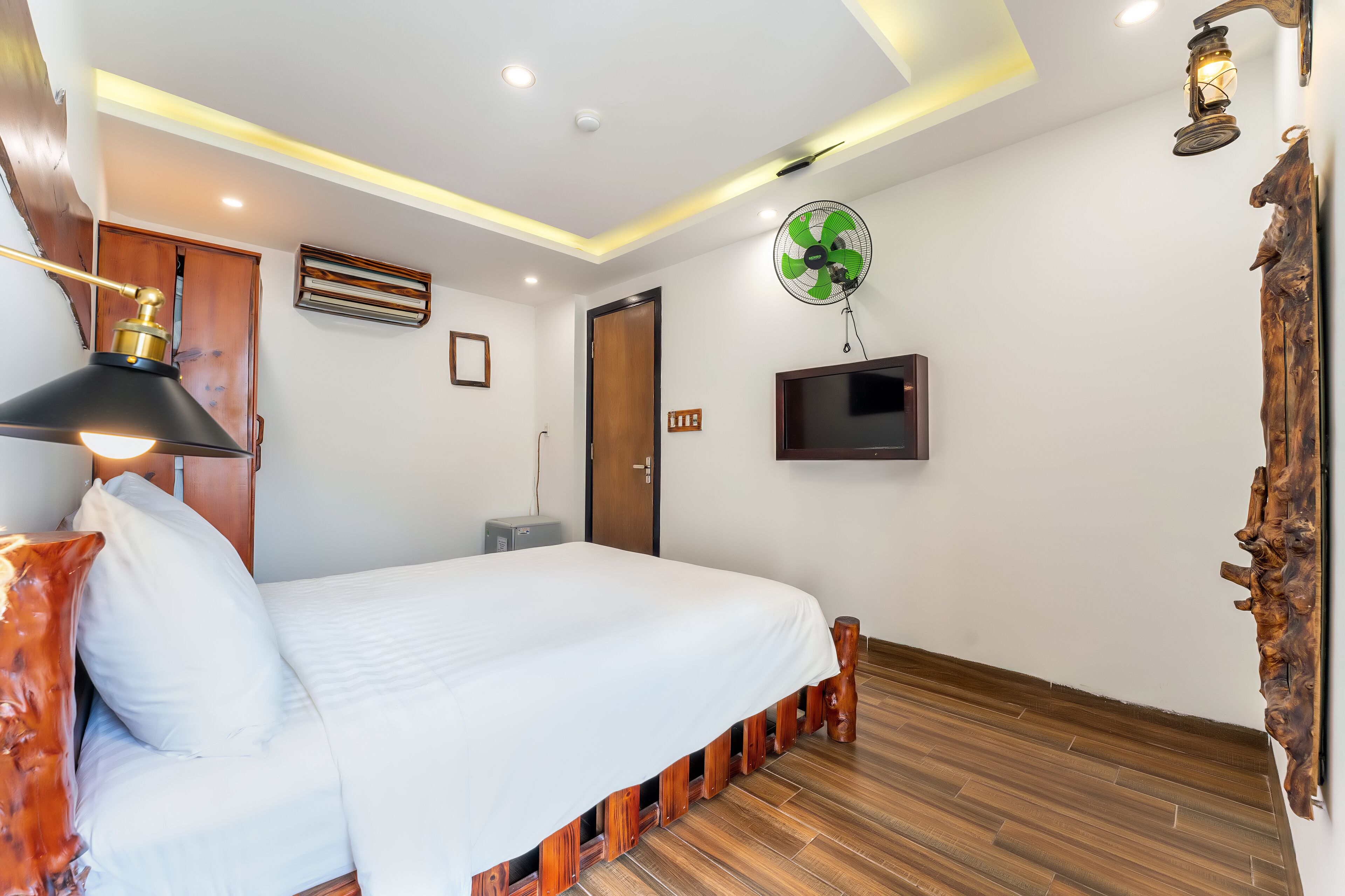 Une chambre à coucher comprenant un lit, un plancher en bois, un ventilateur fixé au mur, une télévision et un luminaire au plafond.