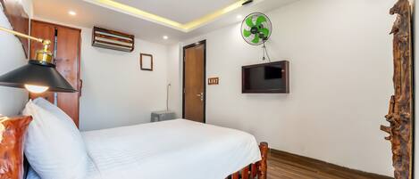 Un dormitorio con cama, suelo de madera, ventilador de pared, televisor y lámpara en el techo.