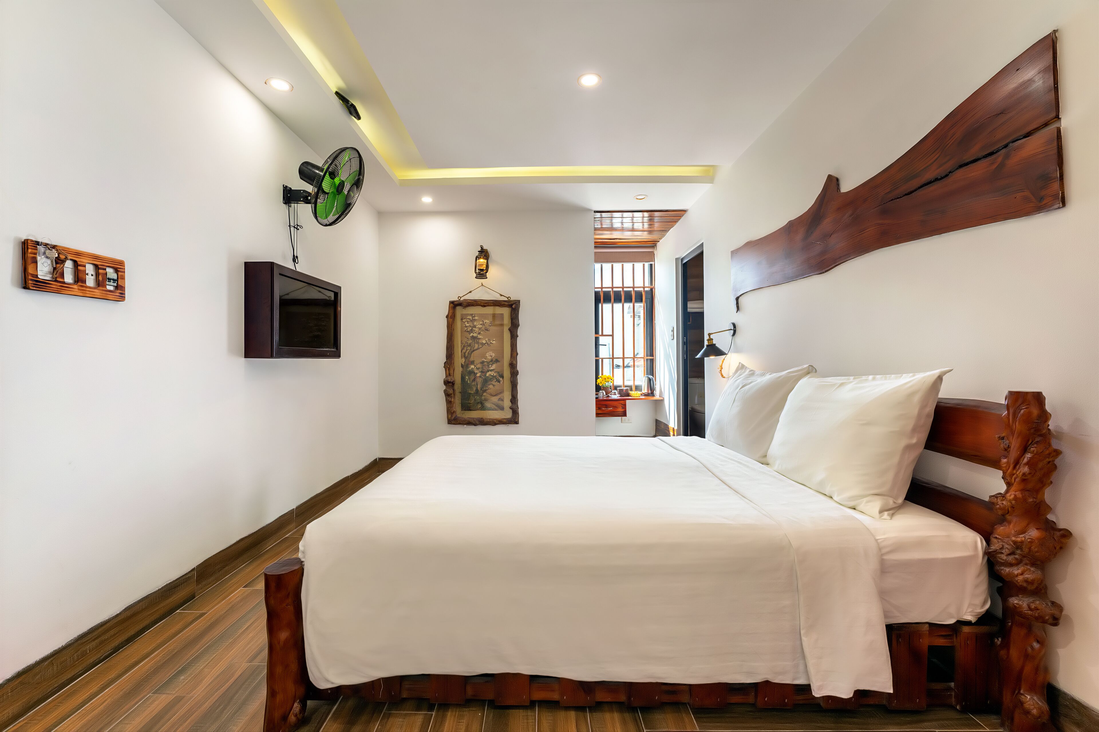 A bedroom with a wooden bed, a wall-mounted fan, a framed artwork, and a ceiling light fixture.