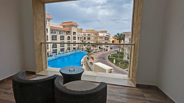 Un balcon agrémenté de chaises en osier et d’une petite table, d’où l’on peut admirer une piscine et un complexe hôtelier avec ses bâtiments et ses palmiers.