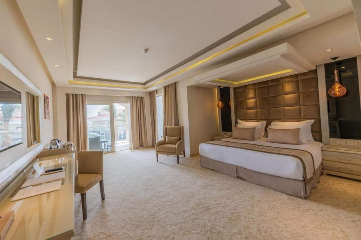 A hotel room with a large bed, a desk, a chair, and a balcony with curtains.