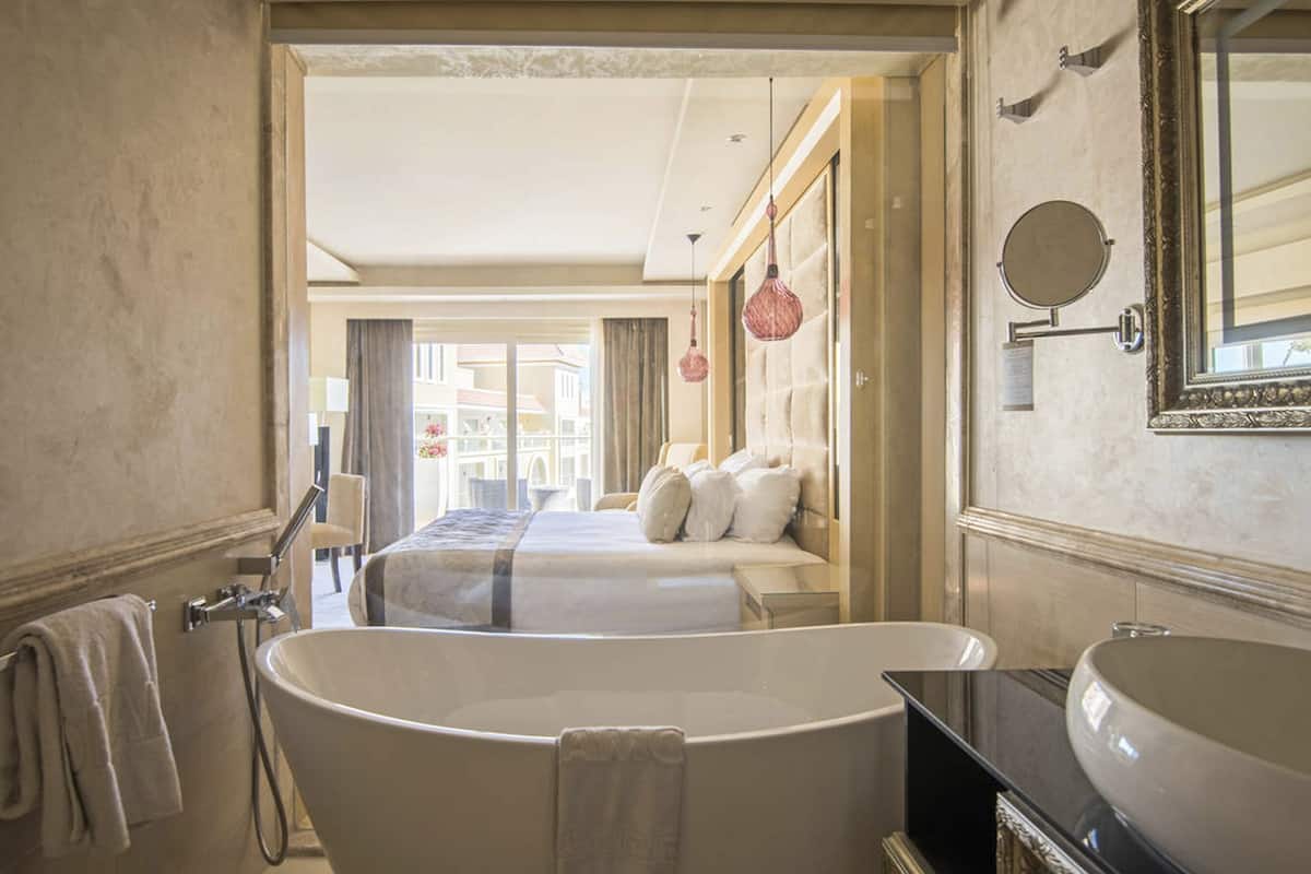 A hotel room with a large bathtub, a bed, a mirror, and a view of the city.