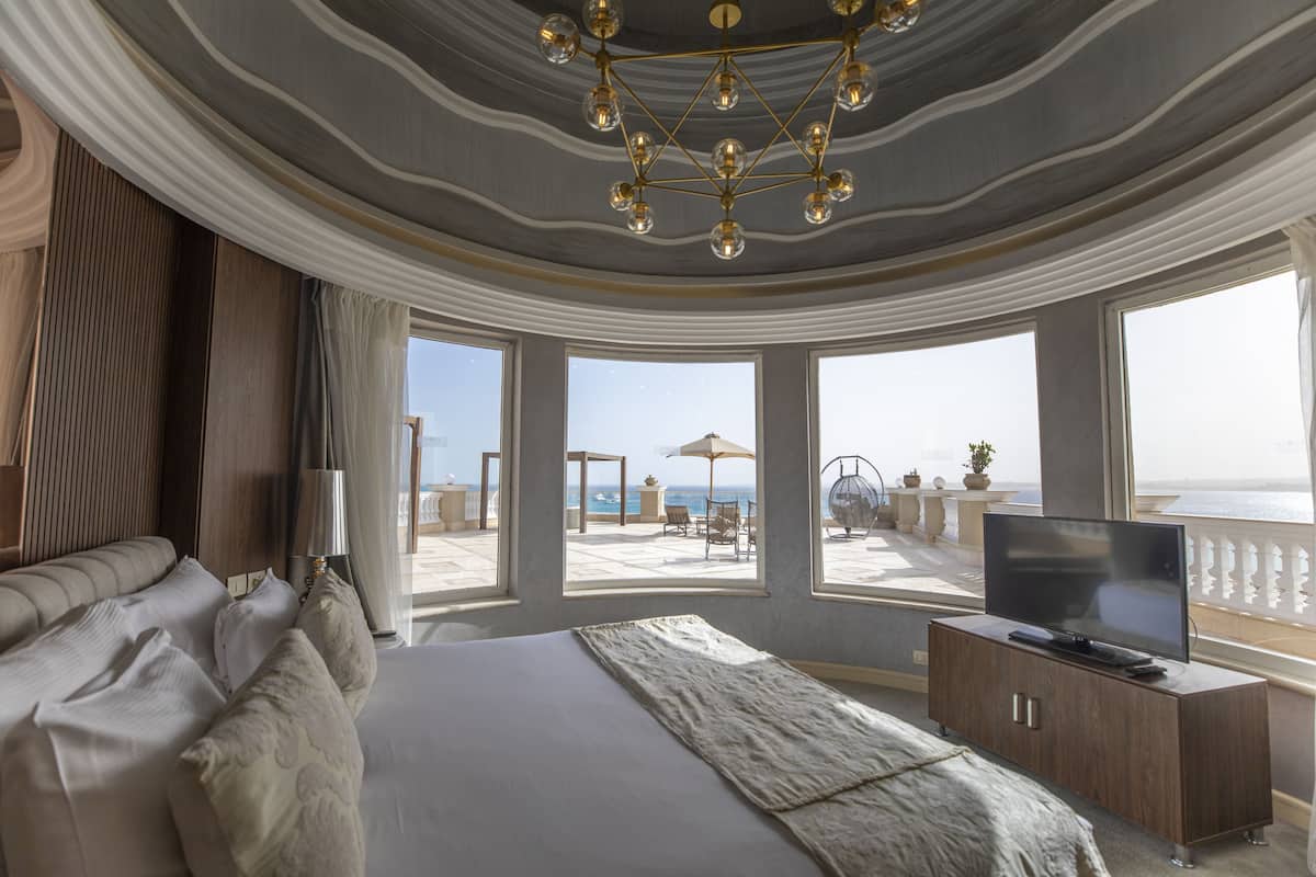 A modern bedroom with a large bed, a television, and a balcony with a view of the sea.