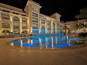 A large, well-lit swimming pool surrounded by a multi-story hotel with balconies and outdoor seating areas. - KaiSol Romance Resort (Sahl Hasheeh)