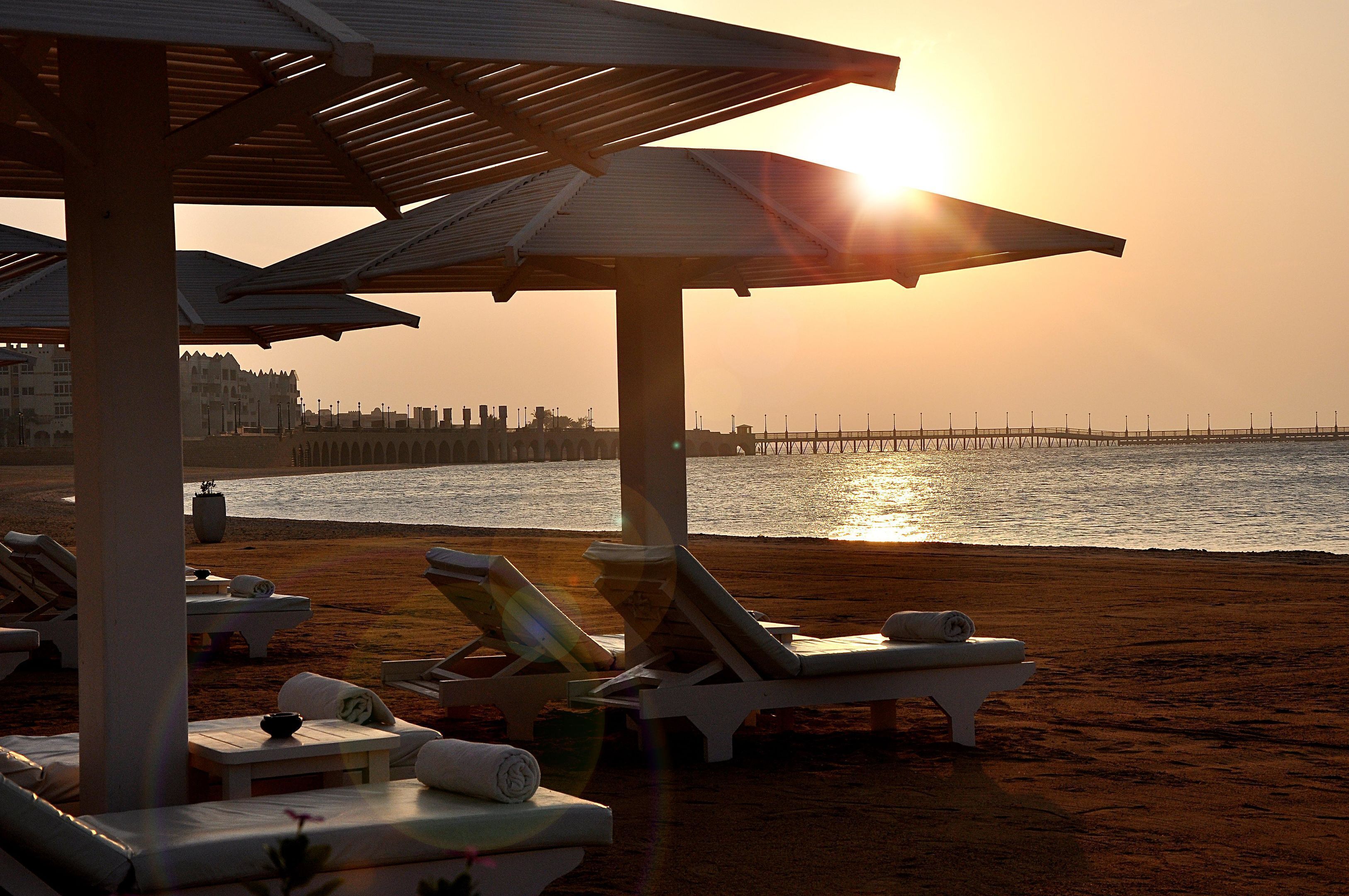 A beachside lounge area with sun loungers and umbrellas, overlooking the sea and a pier in the background.