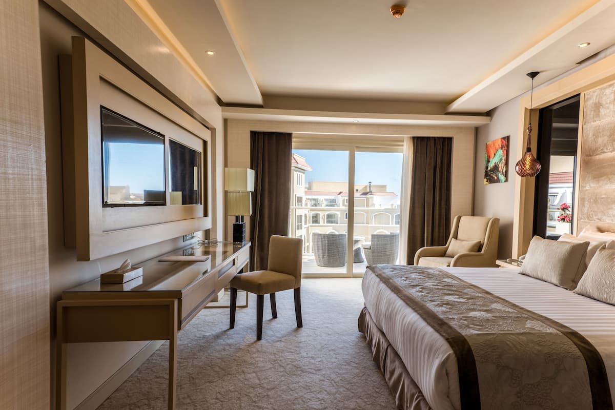 A hotel room with a large bed, a desk with a flat-screen TV, a chair, and a view of the city through large windows.