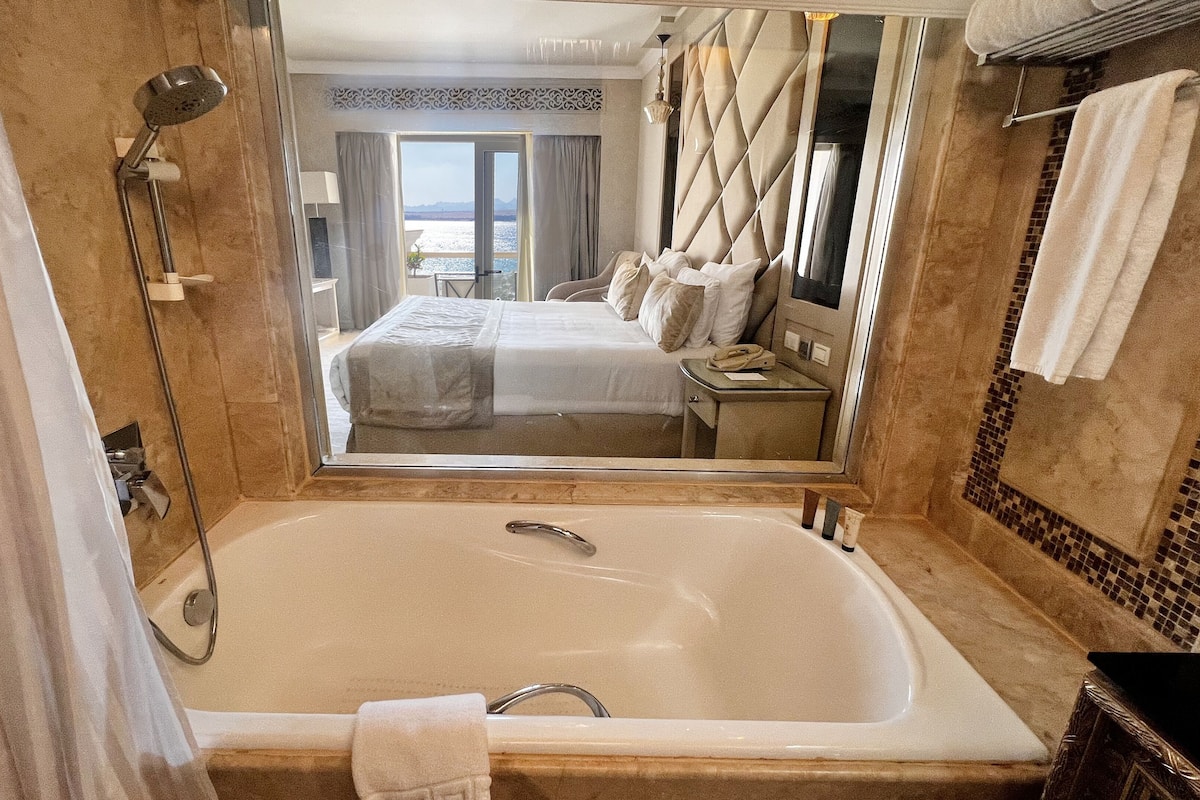 A bathroom with a large bathtub, a mirror, and a view of a bedroom with a bed and curtains.