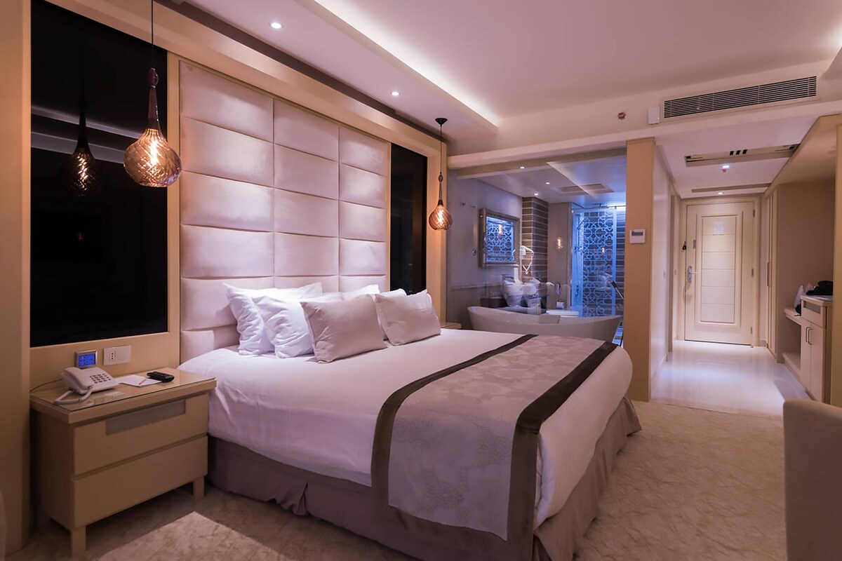 A modern hotel room with a large bed, bedside tables, a desk, and a bathroom visible through an open door.
