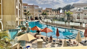 A pool area with lounge chairs and umbrellas, surrounded by palm trees and a building with arched windows. - KaiSol Romance Resort (Sahl Hasheeh)