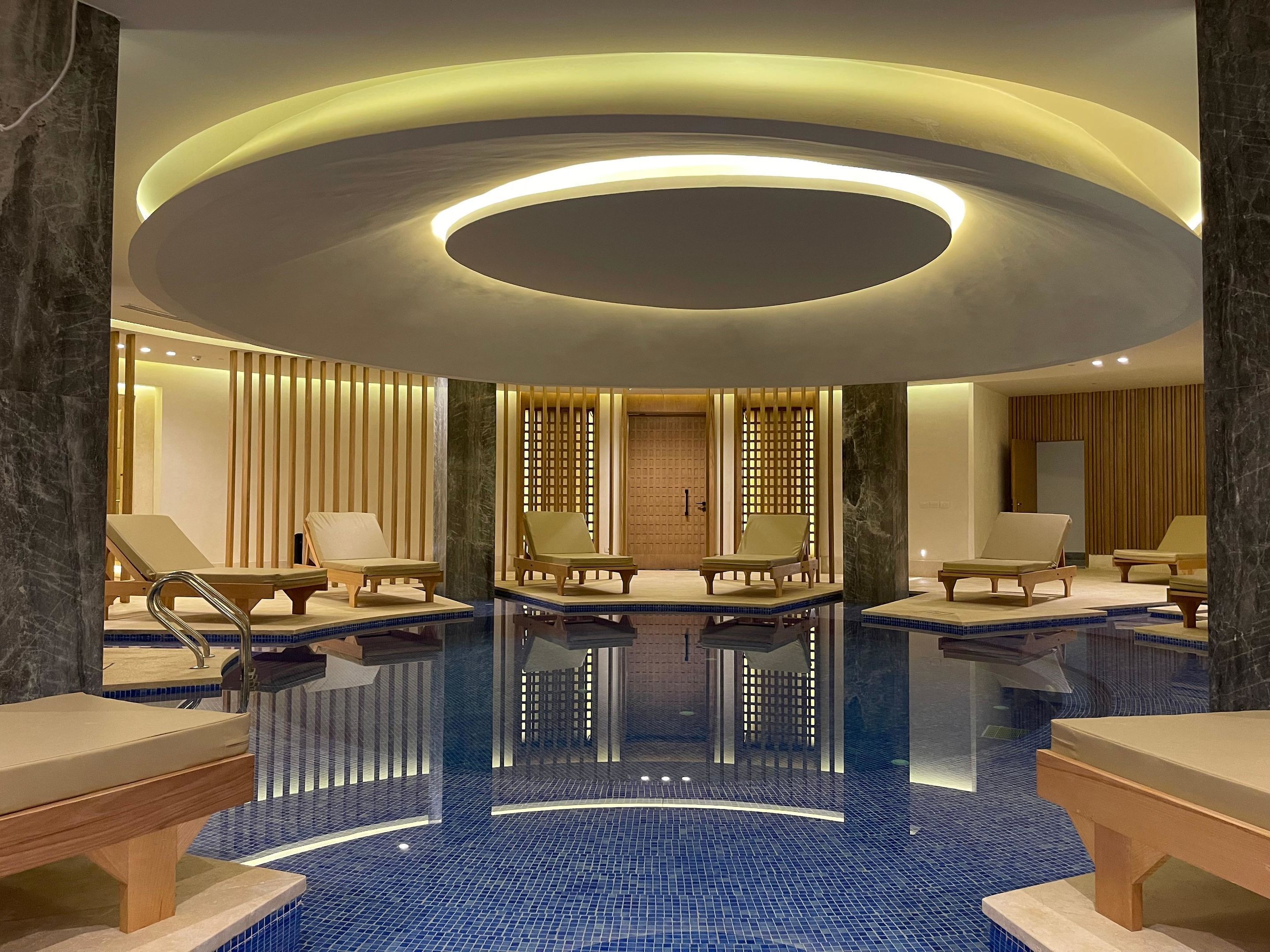 Un spa moderno con piscina central, tumbonas de madera y una lámpara de techo circular.