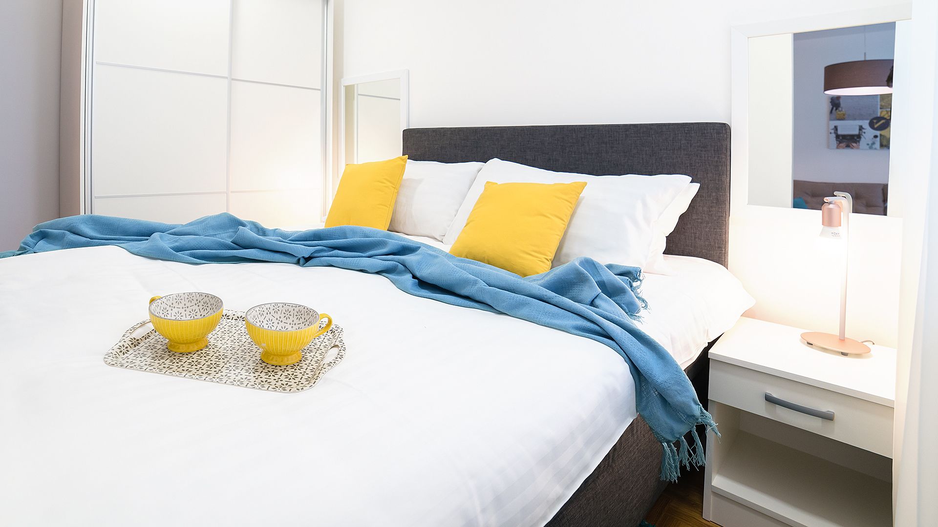 A neatly made bed with a blue blanket, two yellow pillows, and a tray with two yellow cups.