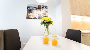 A dining table with two glass mugs of orange juice, a vase with yellow flowers, and a wall-mounted photo collage.