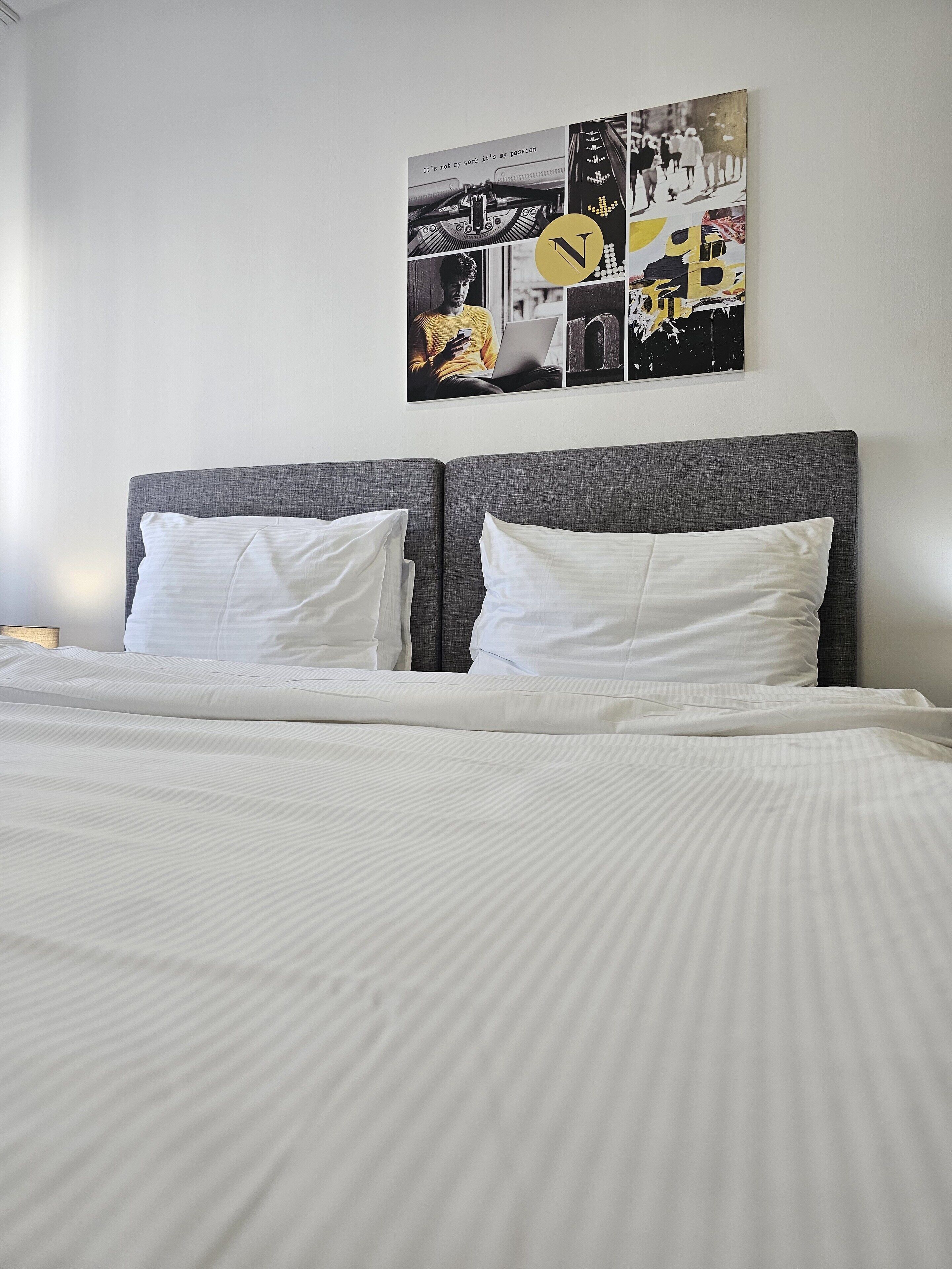 A neatly made bed with white linens and pillows, a grey upholstered headboard, and a wall-mounted artwork depicting various scenes.