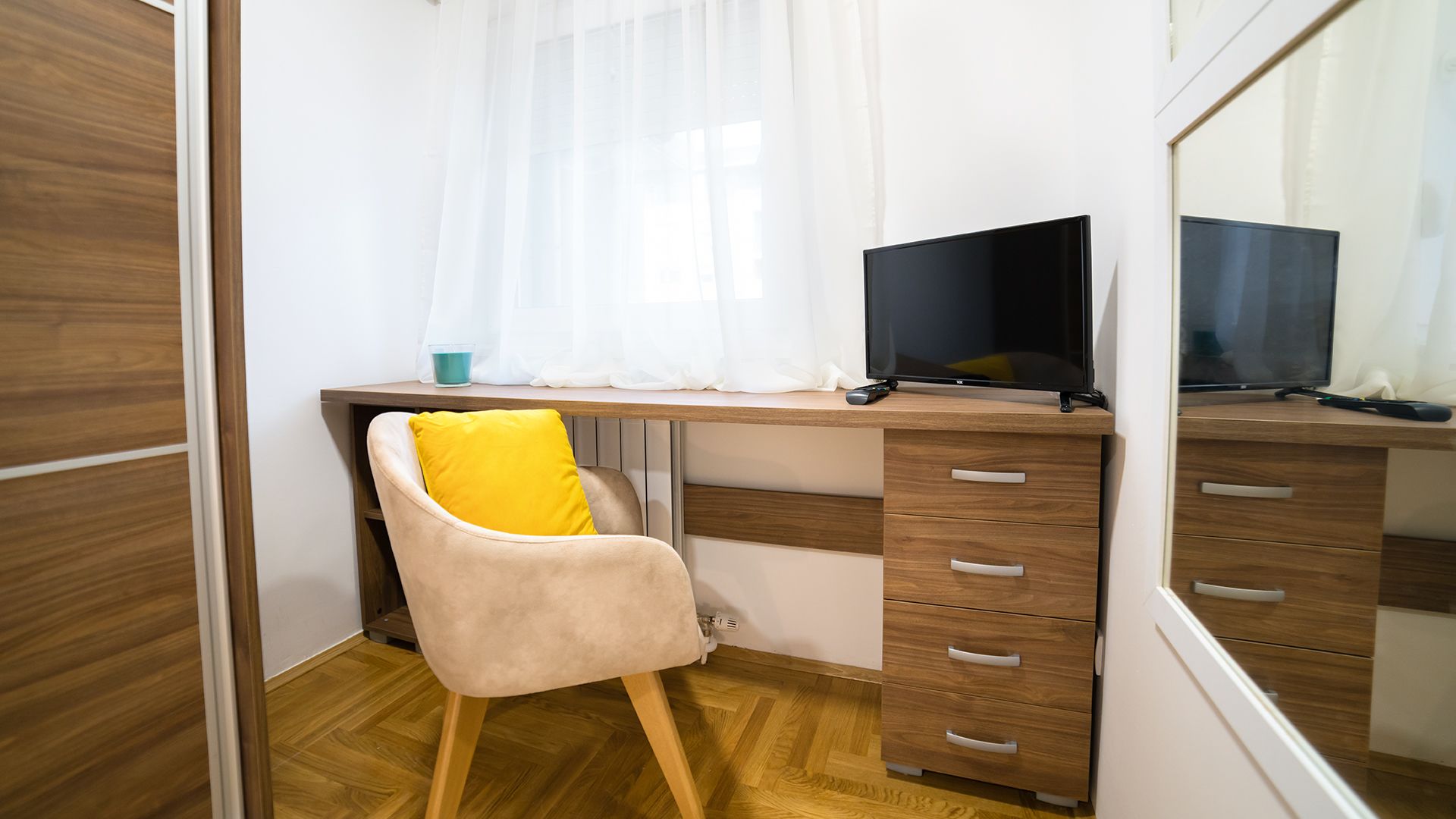 A bedroom with a wooden desk, a chair with a yellow cushion, a TV, and a wardrobe.