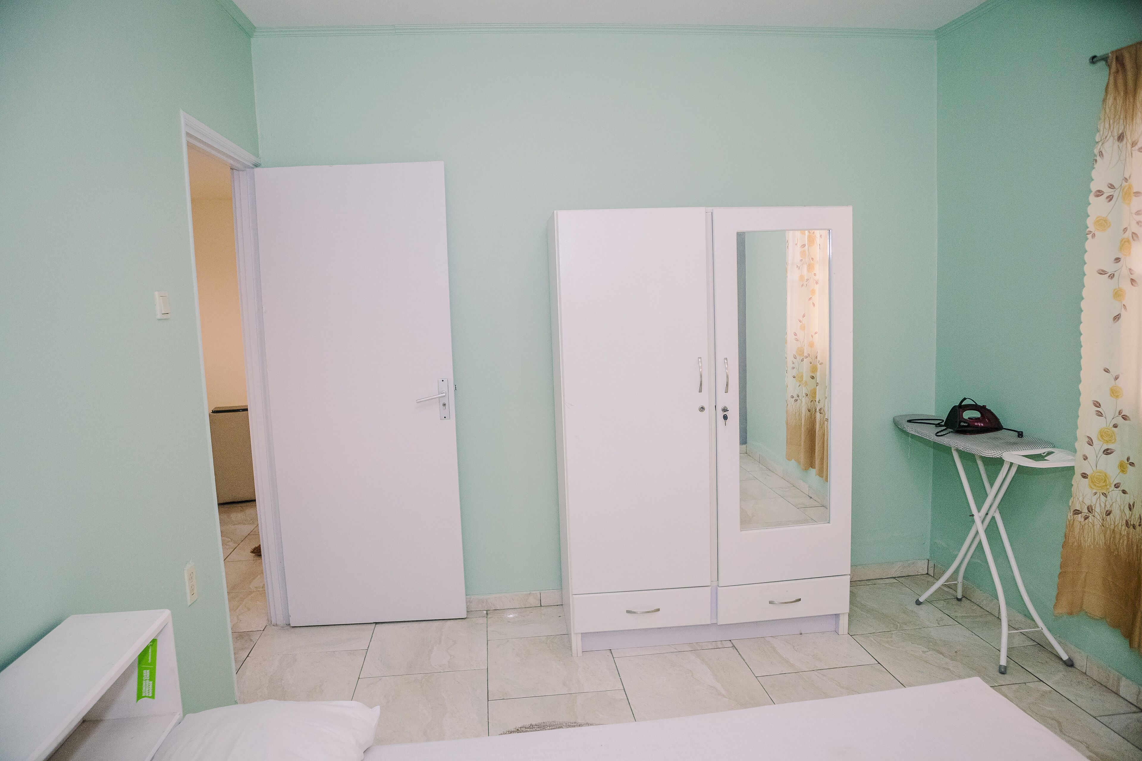 A bedroom with a bed, a wardrobe, a mirror, and a small table with an ironing board.