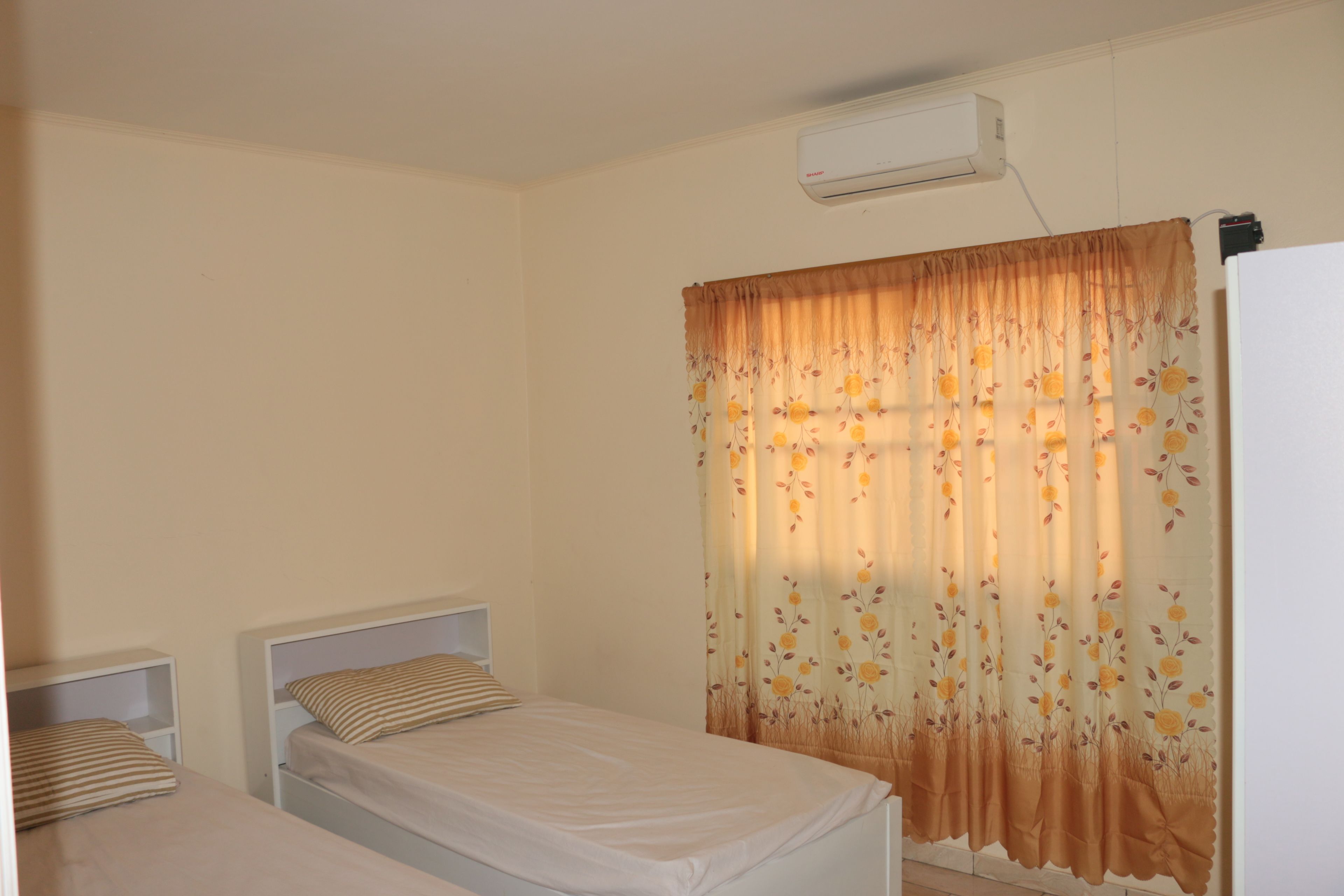 Una habitación pequeña y limpia con dos camas individuales, aire acondicionado blanco y una ventana con cortinas florales.