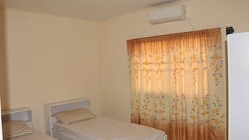 Una habitación pequeña y limpia con dos camas individuales, aire acondicionado blanco y una ventana con cortinas florales.