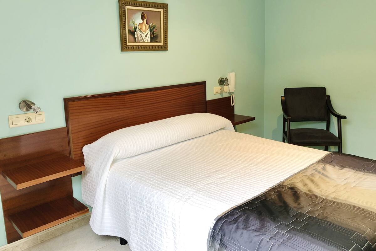 Una cama bien hecha con edredón blanco y manta estampada en negro y gris. Se observa un cabecero de madera, una mesita de noche con una lámpara y un cuadro enmarcado en la pared. Una silla negra está colocada junto a la pared.